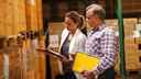 Man and woman with clipboards inspecting boxed products on pallets in a warehouse