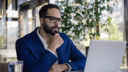 Man wearing suit, sitting in front of laptop, looking quizzical 