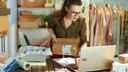 Businesswoman handling packaging for e-commerce while working on laptop, against a background of a small clothing business office