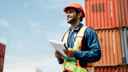 A customs worker wearing a high-viz jacket standing in front of shipping containers and carrying a clipboard with customs documentation